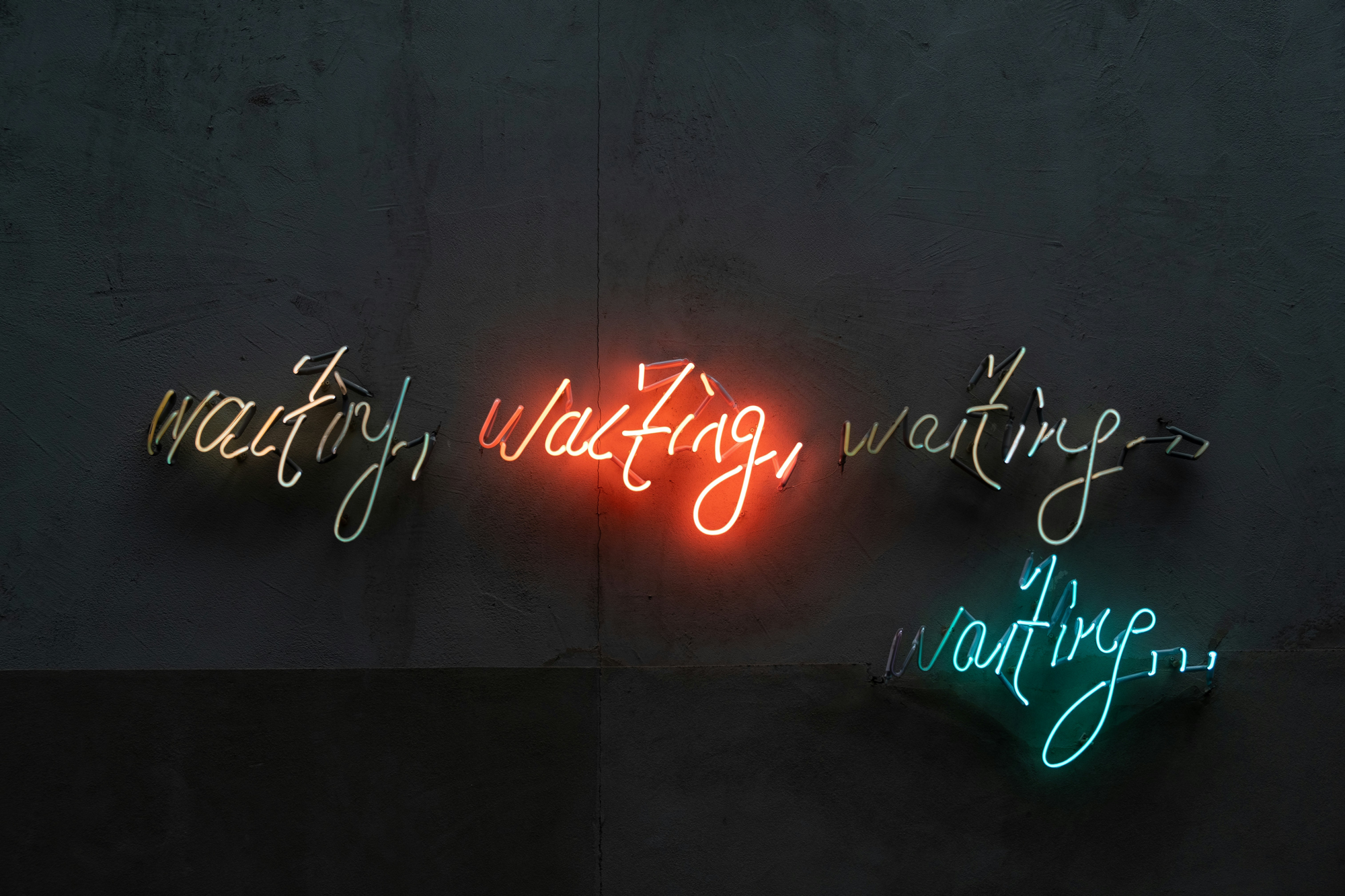 Neon signs on a dark wall spelling 'waiting, waiting, waiting, waiting' in different colors.